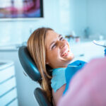Blonde woman smiles while sitting in a dental chair looking at her dental hygienist holding a dental mirror